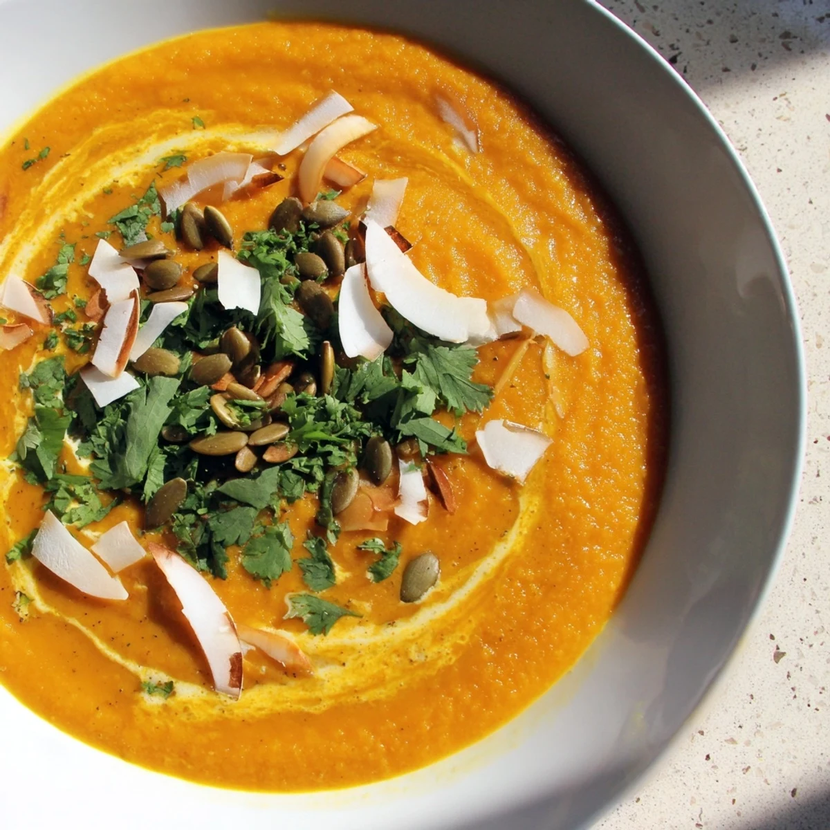 A warm bowl of Roasted Carrot and Ginger Soup with Coconut Milk, topped with toasted coconut flakes and pumpkin seeds for crunch.