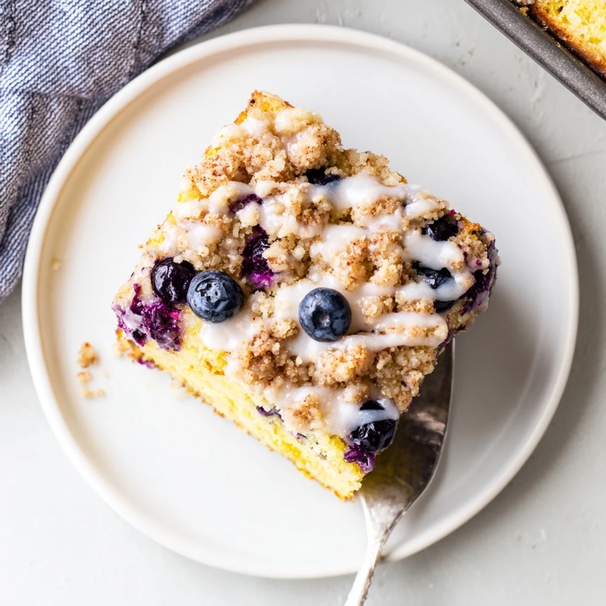 A slice of moist Lemon Blueberry Coffee Cake with a golden crumb topping and juicy blueberries peeking through, served on a white plate.
