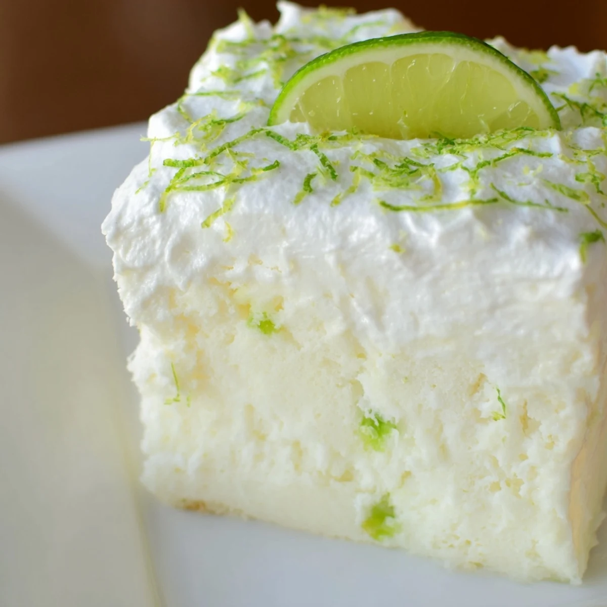 A slice of lime poke cake with whipped topping sits on a white plate, featuring moist cake soaked in bright green lime gelatin and crowned with fluffy cream and fresh zest.