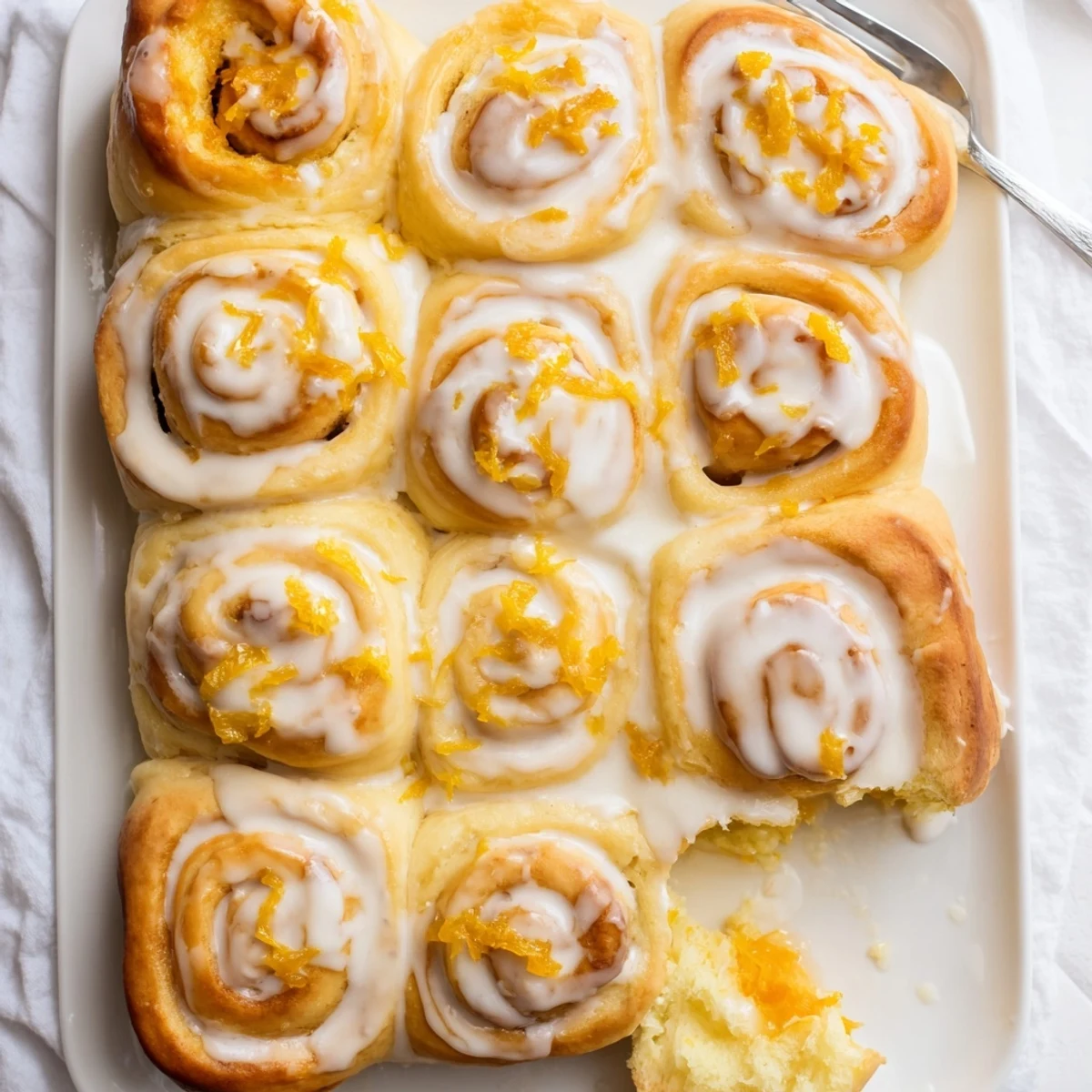 A close-up of golden-brown Homemade Glazed Orange Rolls drizzled with a sweet citrus glaze, showcasing their soft, fluffy texture in a white ceramic baking dish.