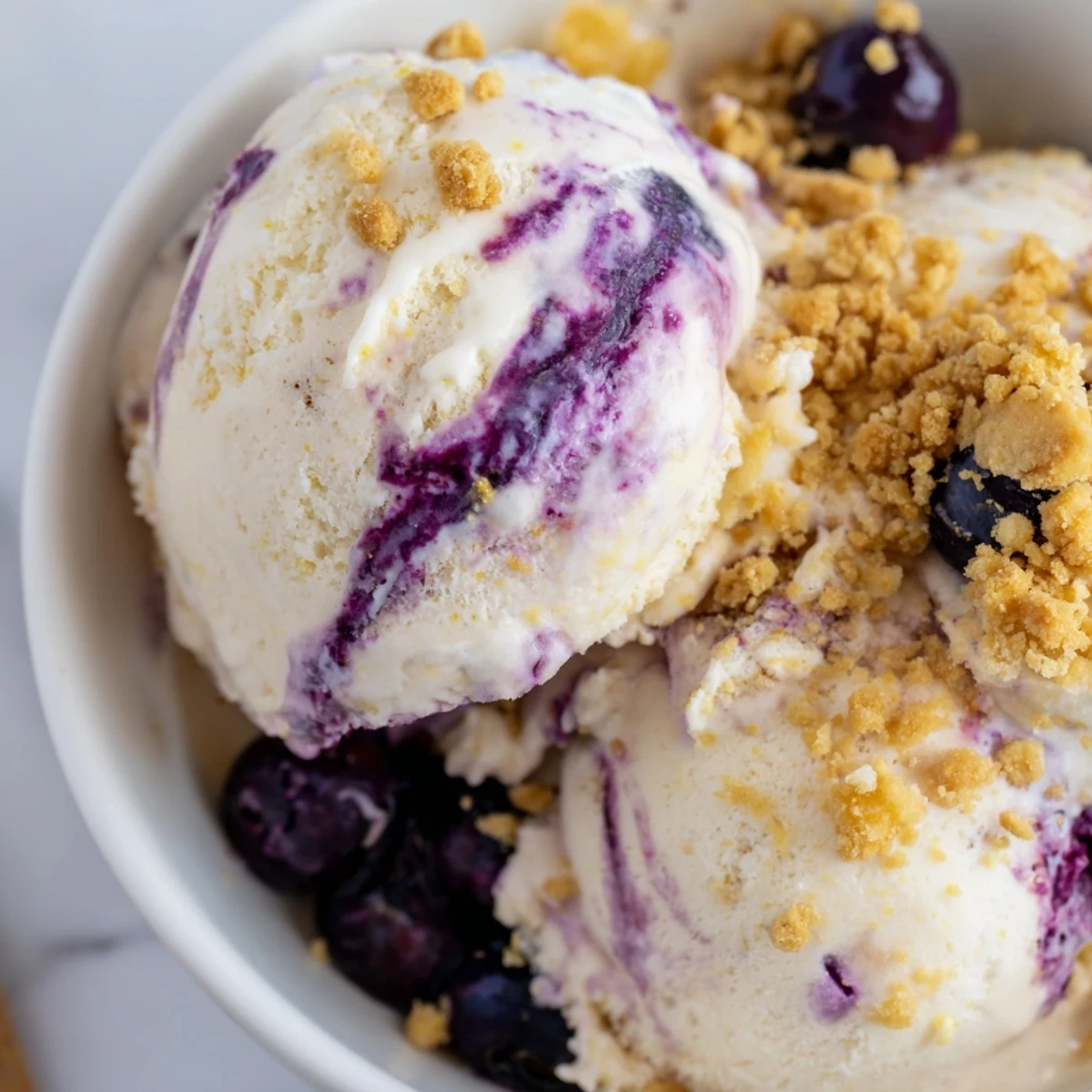Creamy Lemon Blueberry Cheesecake Ice Cream in a scoop, topped with fresh berries and graham crumbs on a rustic table.
