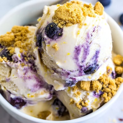 A close-up of vibrant blueberry swirls in homemade Lemon Blueberry Cheesecake Ice Cream, melting slightly beside a crisp lemon shortbread cookie.